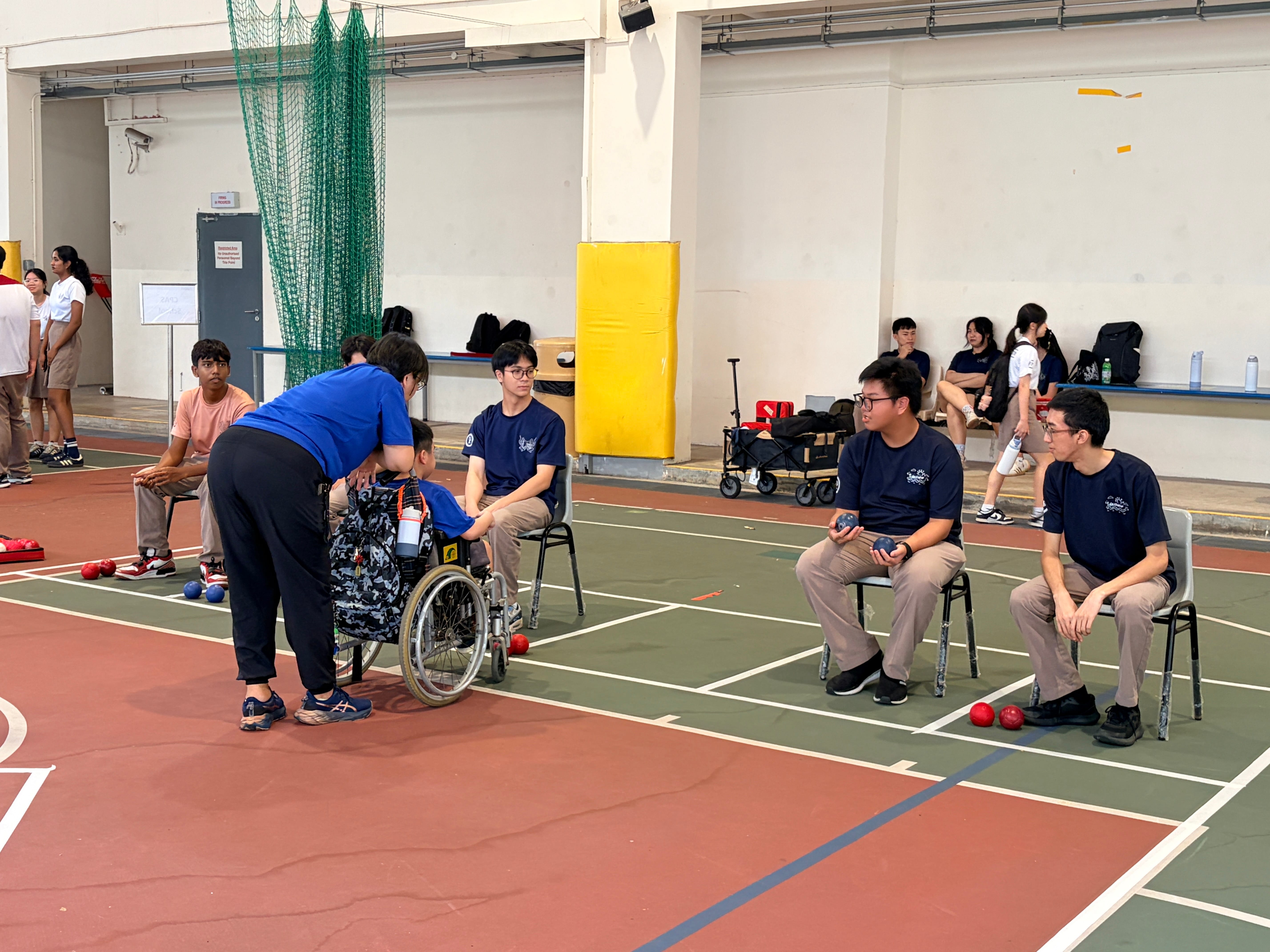 Students seated on a sports court playing Boccia, with one student in a wheelchair being assisted by a facilitator in a blue shirt.